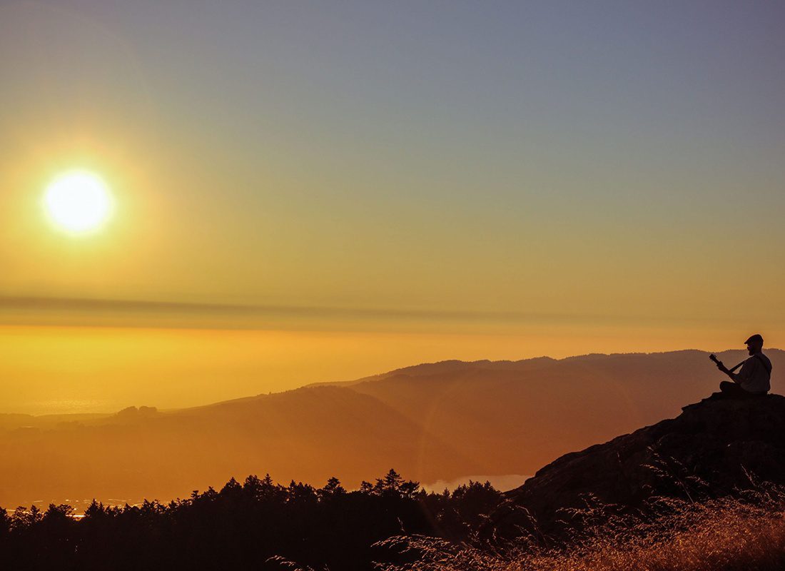 Contact - Silhouette of a Man Sitting Down at the Top of a Grassy Hill Playing a Guitar While Looking at Distance Mountain Ranges During an Orange-toned Sunrise